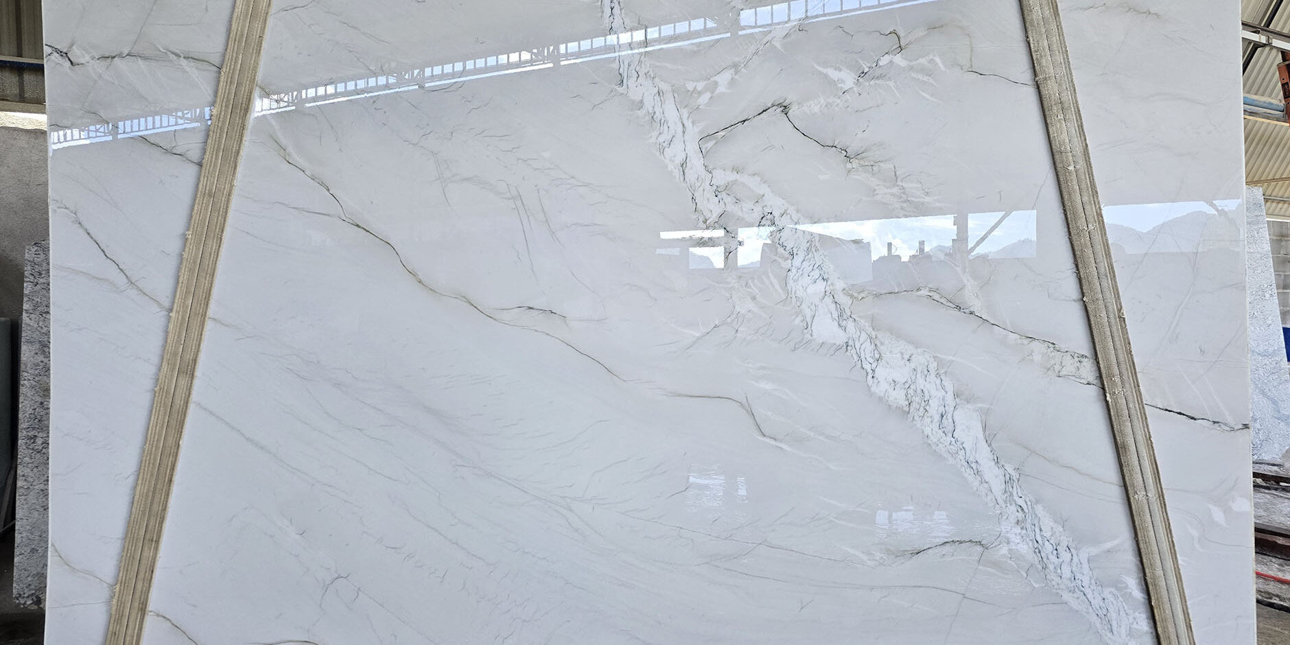 A large, polished slab of Bianco Superiore Quartzite, showing its distinct crystalline texture and elegant grey and brown veining, displayed vertically on wooden supports.