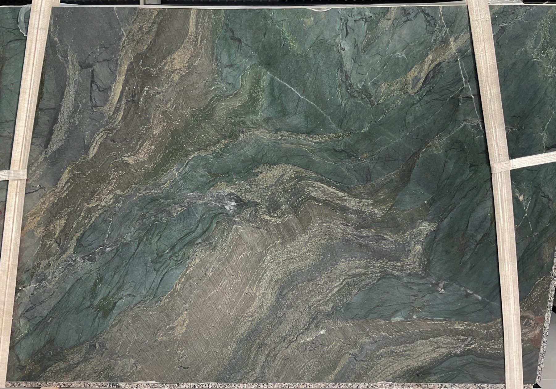 A full view of a Sombori Crystal quartzite slab suspended on lifting straps, displaying its deep emerald green background, dark forest undertones, and dramatic gold veining.