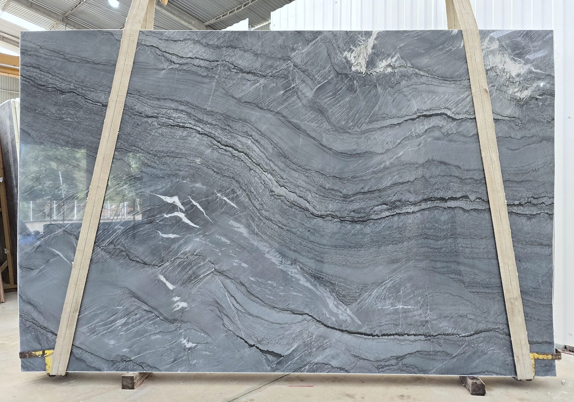 A full view of a leathered Platinum Blue quartzite slab suspended on lifting straps, displaying its steel-blue background, deep charcoal waves, and crystal veins.